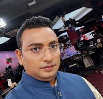Portrait of a man wearing glasses and a blue traditional outfit in a TV studio with cameras and lights in the background.