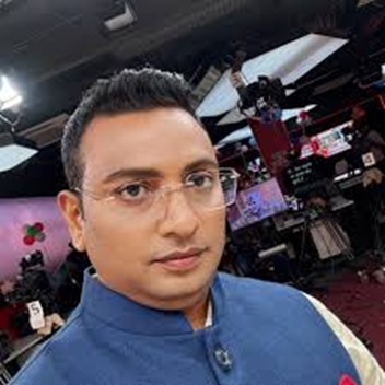 Portrait of a man wearing glasses and a blue traditional outfit in a TV studio with cameras and lights in the background.