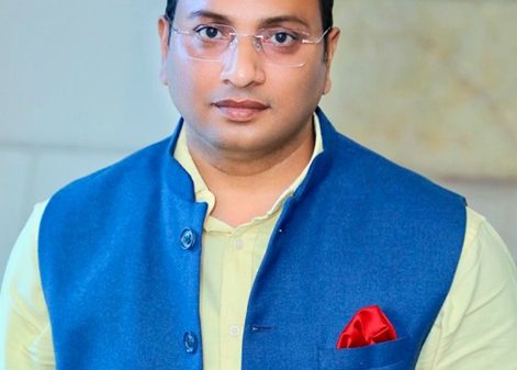 Portrait of a man wearing a blue waistcoat over a pale yellow shirt, with a red pocket square and glasses, looking at the camera in an indoor setting.