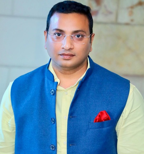 Portrait of a man wearing a blue waistcoat over a pale yellow shirt, with a red pocket square and glasses, looking at the camera in an indoor setting.
