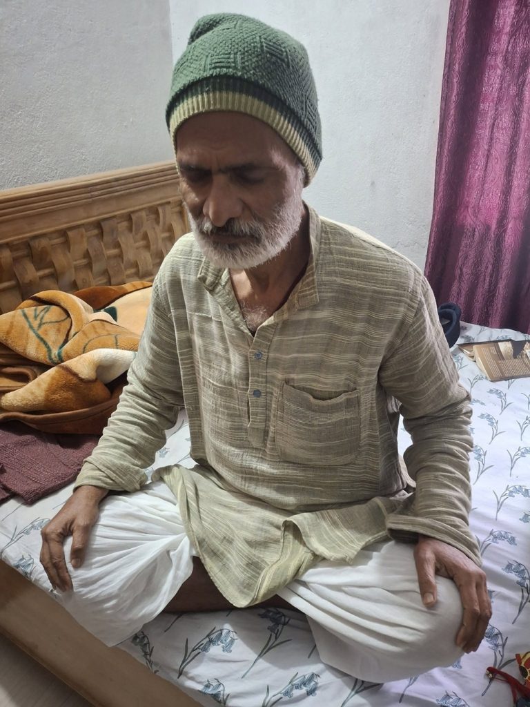 amarnath jha | No. 1 Indian Media News Portal Elderly man with a green knitted cap and white beard sits cross-legged on a bed, looking down in a sunlit room.
