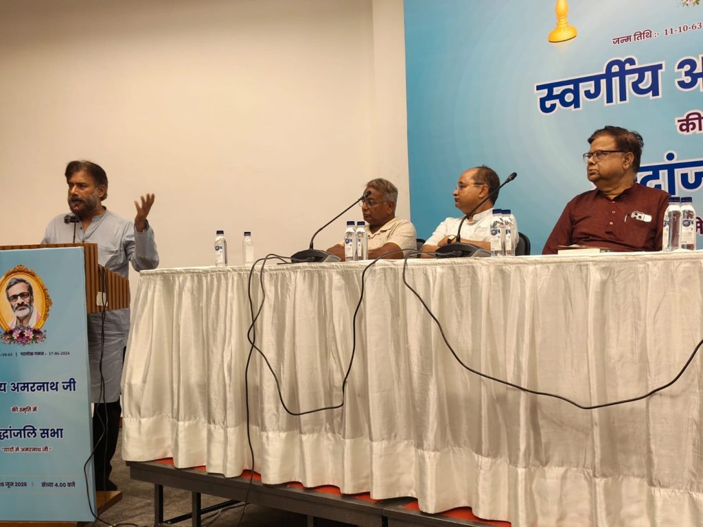 Speaker at a podium addressing a panel, with three men seated at a long table on a stage backdrop in blue text signage and water bottles nearby