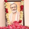 Framed portrait of a bearded man with glasses, in traditional attire, decorated with red and white flower garlands at a shrine.