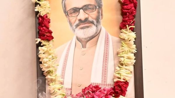 Framed portrait of a bearded man with glasses, in traditional attire, decorated with red and white flower garlands at a shrine.