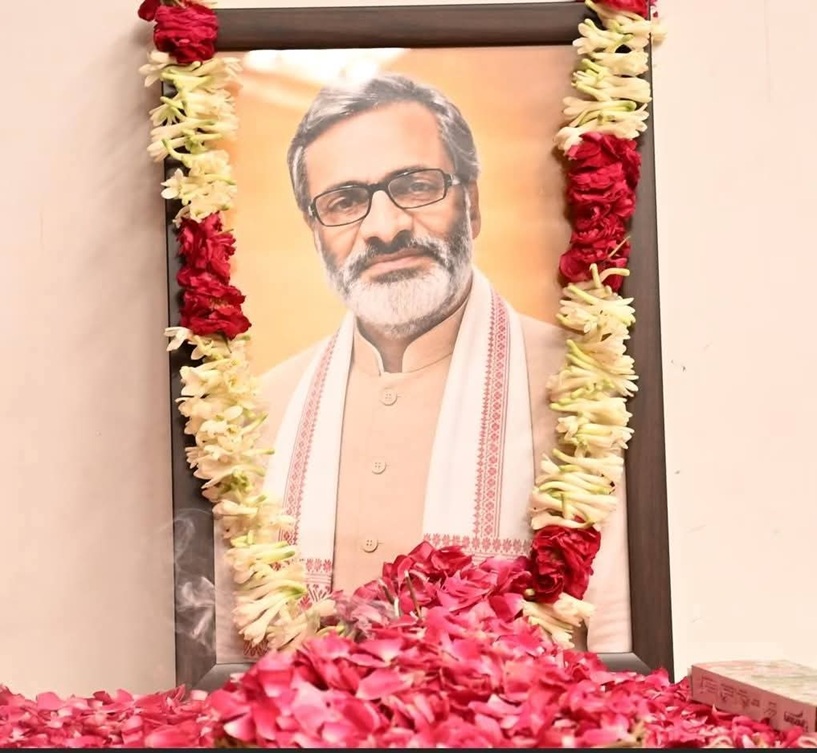Framed portrait of a bearded man with glasses, in traditional attire, decorated with red and white flower garlands at a shrine.