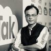 Man wearing glasses and a striped shirt with a dark vest stands with arms crossed in front of a wall featuring a large 'talk' graphic and news icons.