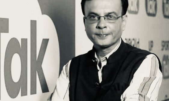 Man wearing glasses and a striped shirt with a dark vest stands with arms crossed in front of a wall featuring a large 'talk' graphic and news icons.