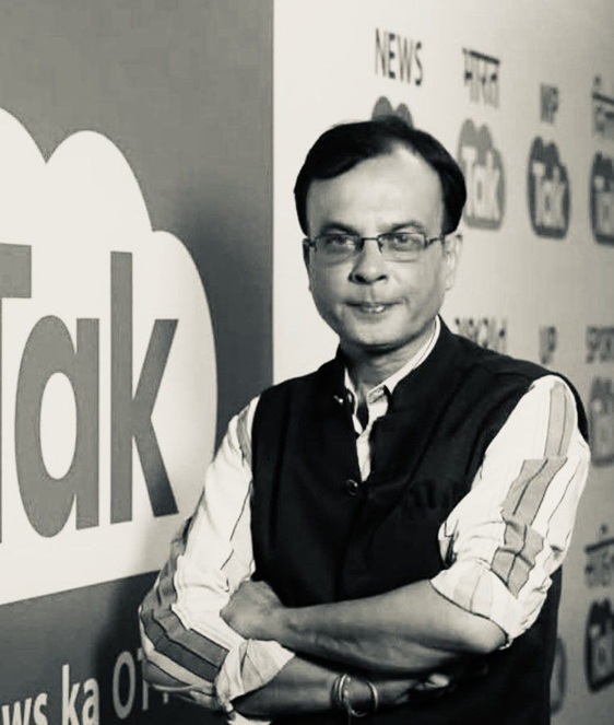 Man wearing glasses and a striped shirt with a dark vest stands with arms crossed in front of a wall featuring a large 'talk' graphic and news icons.