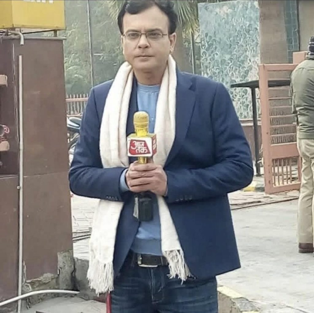 Screenshot | No. 1 Indian Media News Portal Man wearing a blue blazer and white scarf, holding a yellow microphone with a red logo, standing outdoors as a news reporter.