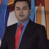 Professional portrait of a man in a black suit, blue shirt, and red tie, standing against a colorful geometric backdrop.