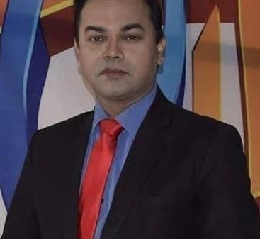 Professional portrait of a man in a black suit, blue shirt, and red tie, standing against a colorful geometric backdrop.