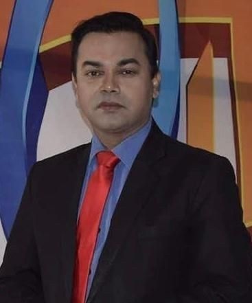 Professional portrait of a man in a black suit, blue shirt, and red tie, standing against a colorful geometric backdrop.