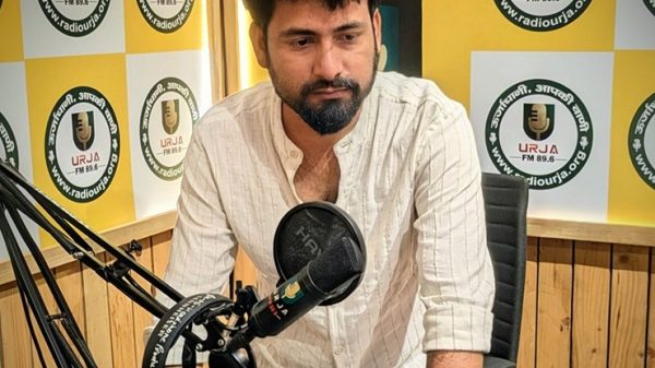 Man adjusting a microphone at a radio studio desk, leaning on a mixing console with headphones nearby and a logoed wall behind him