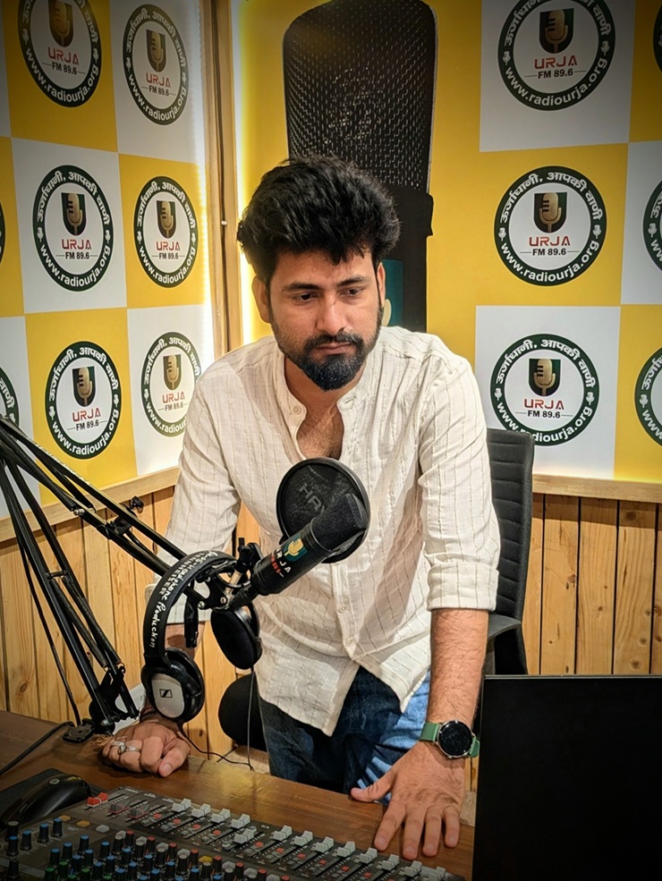 Man adjusting a microphone at a radio studio desk, leaning on a mixing console with headphones nearby and a logoed wall behind him
