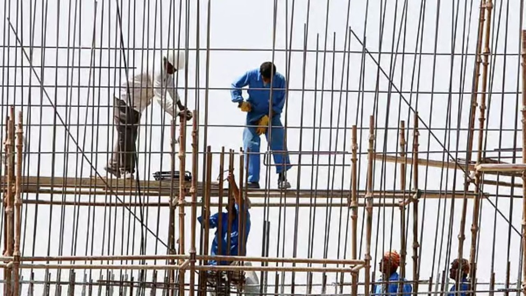 Workers in blue and white coveralls standing on a metal scaffold among vertical rebar during construction, wearing gloves and hard hats (noting safety context).