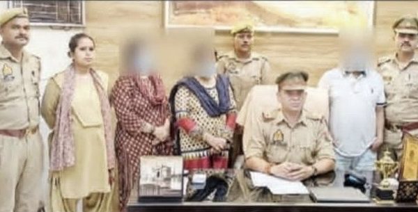 Police officer seated at a desk with papers; several officers and civilians stand behind, faces blurred, in a police station setting with a trophy on the desk.
