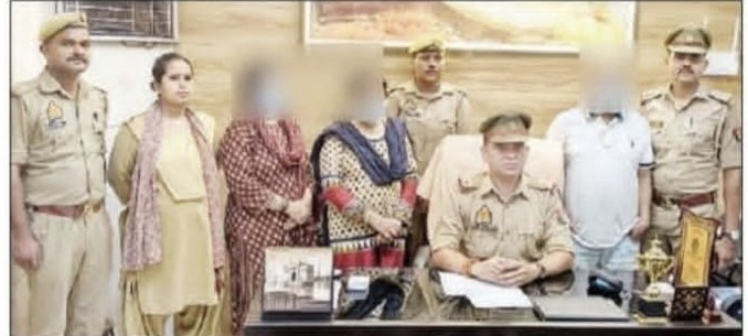 Police officer seated at a desk with papers; several officers and civilians stand behind, faces blurred, in a police station setting with a trophy on the desk.