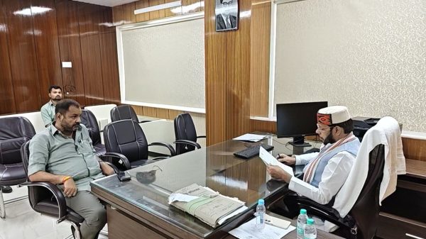 Man in a traditional cap and scarf sits at a glass desk reading a document across from two visitors in a wood-paneled office.