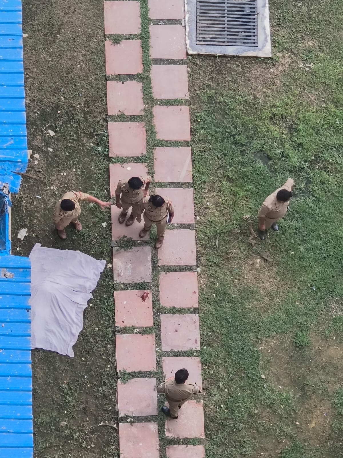 Overhead view of several uniformed people walking along a pink stepping-stone path in a grassy area next to a blue structure and a metal grate nearby.