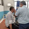 Male patient sits on a hospital bed while a clinician checks his ear in a hospital room with blue walls and equipment nearby.