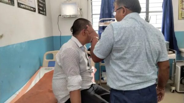 Male patient sits on a hospital bed while a clinician checks his ear in a hospital room with blue walls and equipment nearby.