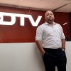 Man in a light dotted shirt stands with hands in pockets in front of a white reception counter at an NDTV office, with a large NDTV sign behind him.