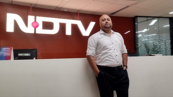 Man in a light dotted shirt stands with hands in pockets in front of a white reception counter at an NDTV office, with a large NDTV sign behind him.
