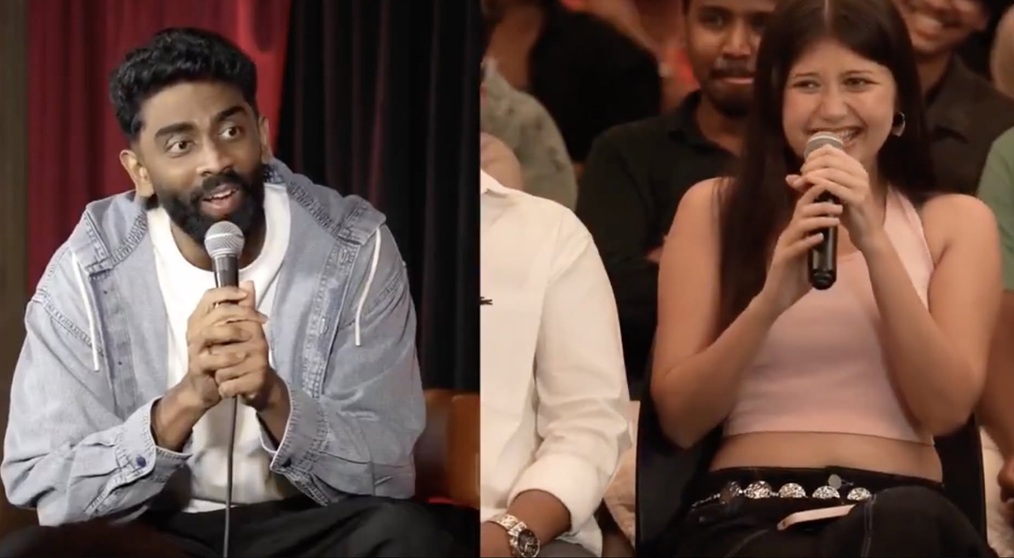 Two people sit side by side on a stage, each holding a microphone and speaking to an audience.