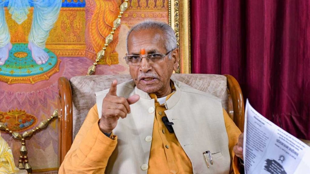 champat | No. 1 Indian Media News Portal Elder Indian man in an orange kurta and beige vest points while speaking, holding a document.