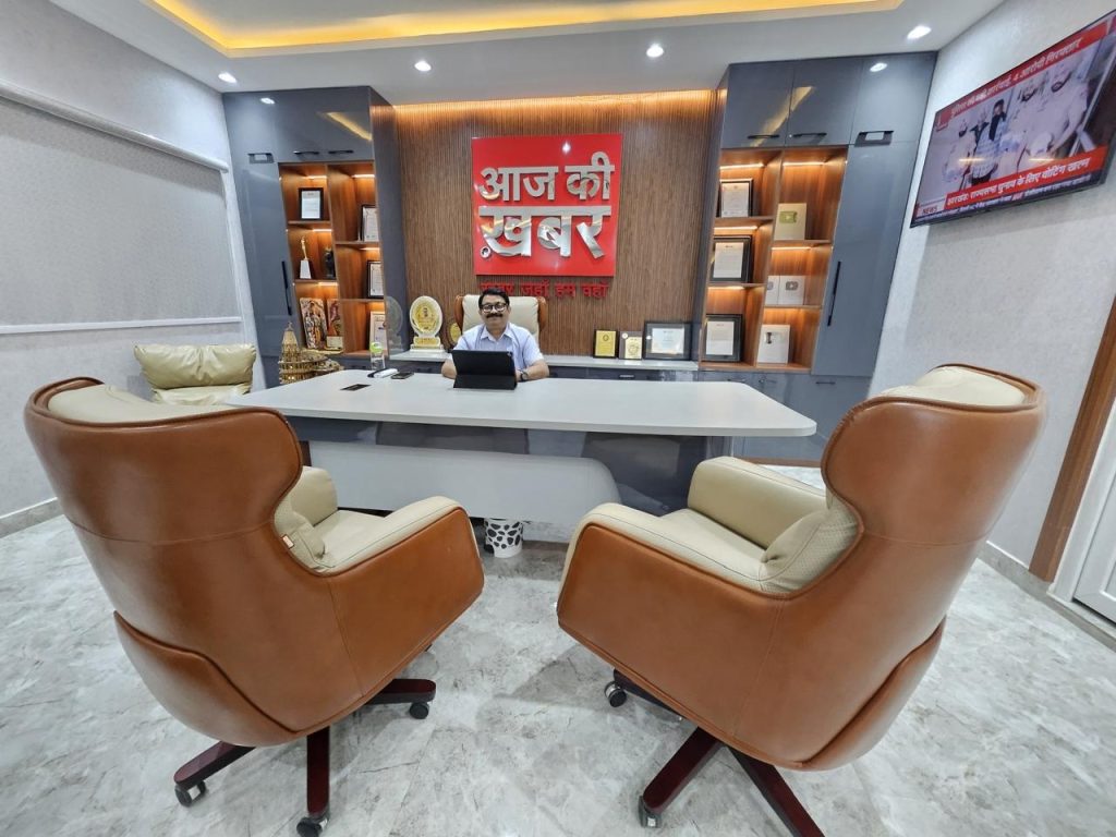 Office reception with a man at a white desk, tan leather chairs, and a red Hindi sign behind him reading 'आज की खबर'.