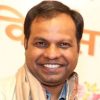 Smiling man with short dark hair wearing a light beige scarf, portrait-style photo with a soft, colorful background.