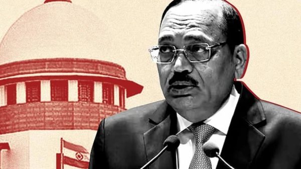 A suited man with glasses speaks into two microphones at a formal event, with a red-tinted government building backdrop behind him.