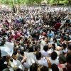 Large crowd of protesters packed together outdoors, many holding up white placards amid green trees in the background.