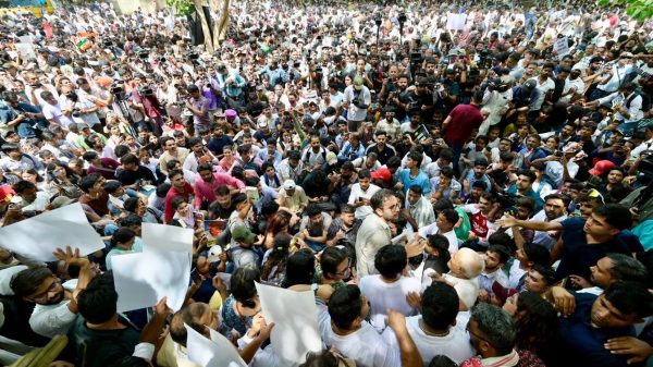 Large crowd of protesters packed together outdoors, many holding up white placards amid green trees in the background.