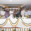 Group of people standing behind a long, decorated table at a formal press event, hands joined in a handshake.