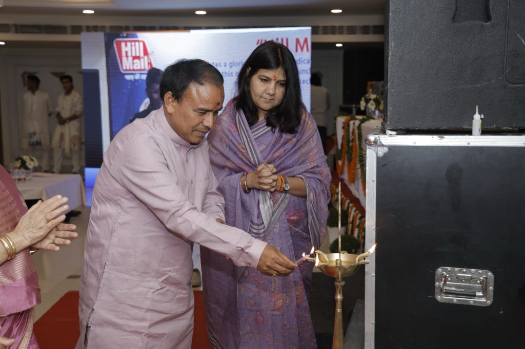 d918405e-33bc-4fb3-b029-007298e682f9 | No. 1 Indian Media News Portal Man in a pink kurta lighting a ceremonial lamp with a woman in a lavender sari watching at an event, Hill Mall banner in background
