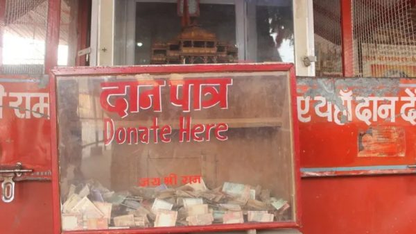 Red donation box with a transparent front showing cash inside; sign reads 'Donate Here' in English and Hindi.