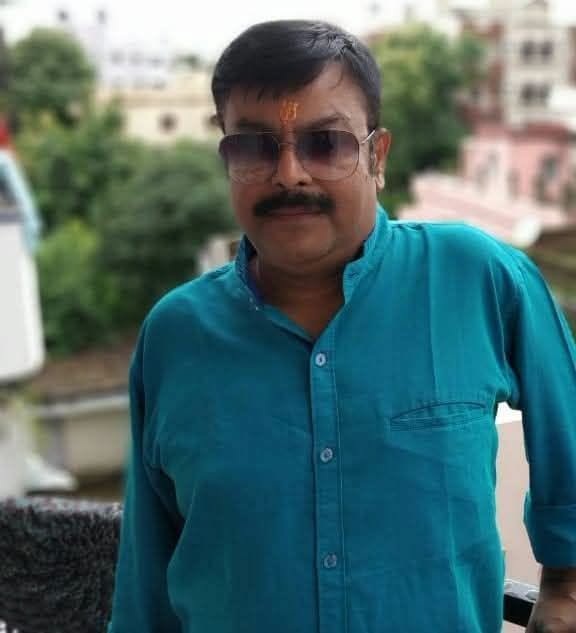 Man with moustache wearing teal shirt and sunglasses on a balcony, forehead with tilak, looking at the camera