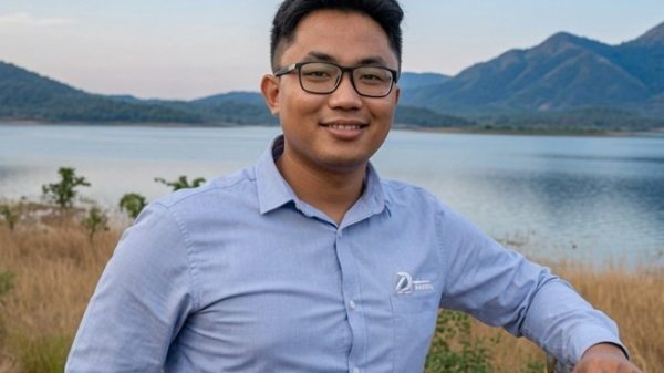 Man wearing glasses and a light blue button-down shirt stands at a railing by a lake with hills in the background, smiling at the camera.