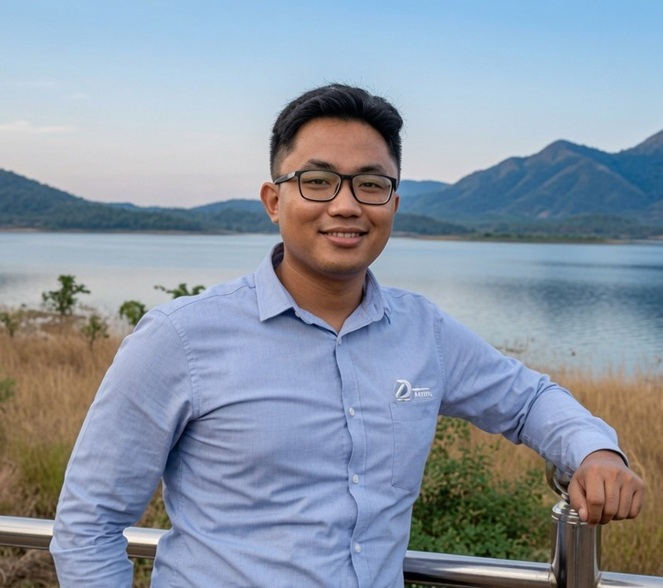 Man wearing glasses and a light blue button-down shirt stands at a railing by a lake with hills in the background, smiling at the camera.