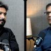 Split-screen interview: left man in black shirt speaking into a microphone, right man in blue shirt with glasses sits before a mic; divider in middle. Typical podcast setup.