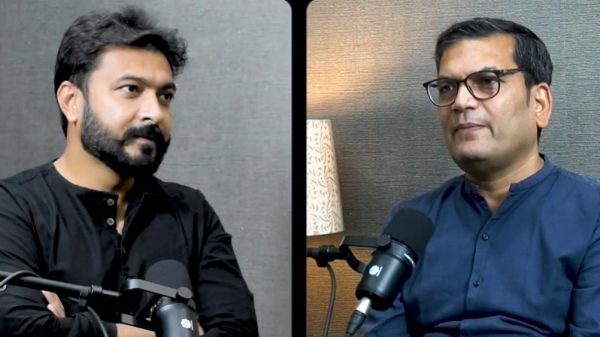 Split-screen interview: left man in black shirt speaking into a microphone, right man in blue shirt with glasses sits before a mic; divider in middle. Typical podcast setup.