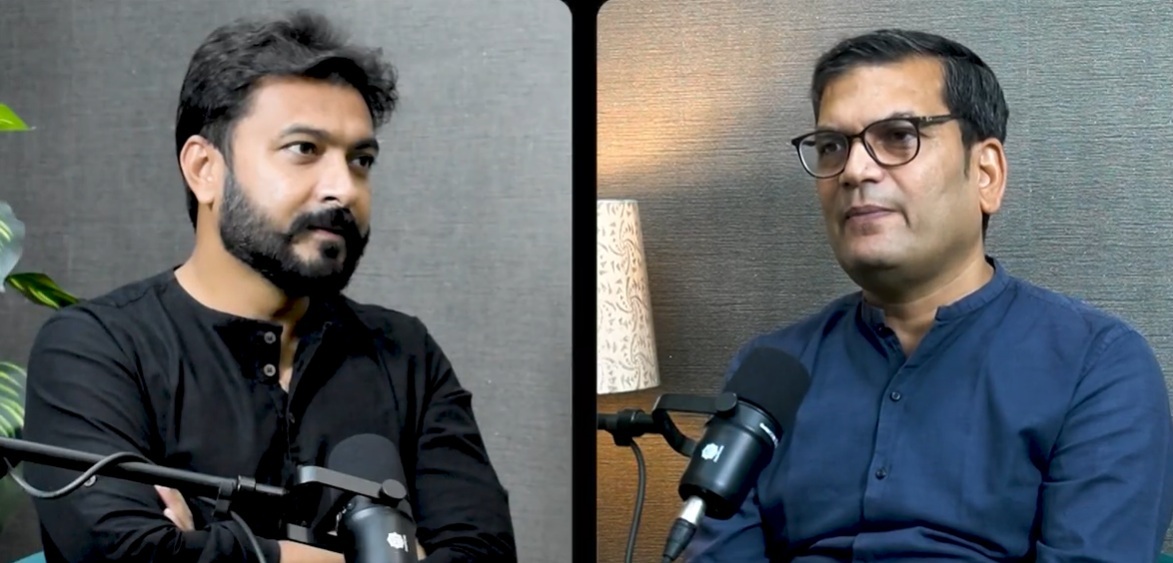 Split-screen interview: left man in black shirt speaking into a microphone, right man in blue shirt with glasses sits before a mic; divider in middle. Typical podcast setup.