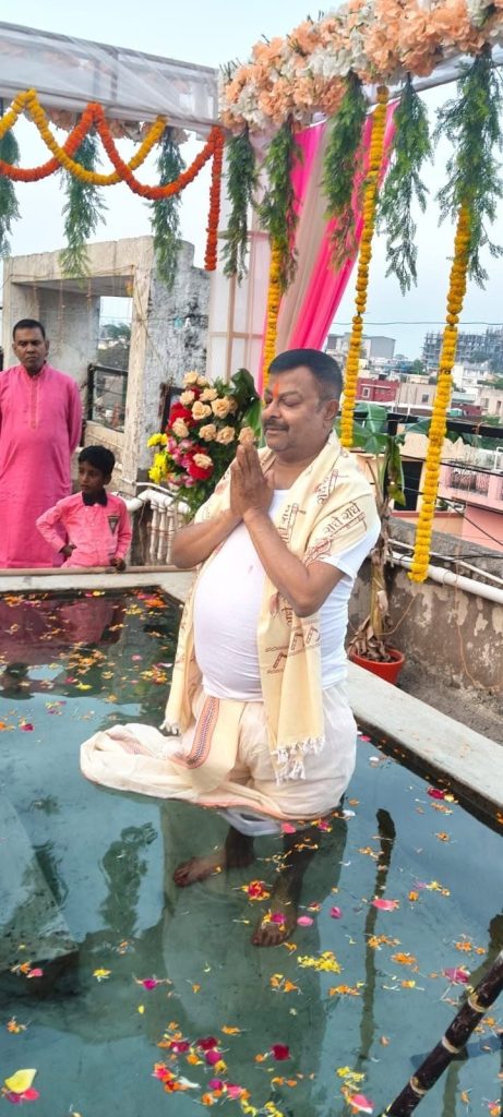 Man in white kneels in a shallow rooftop pool, hands in prayer, during a flower-decorated ceremony with onlookers in pink nearby.
