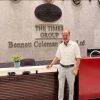 Man in a white shirt and gray pants stands at a reception desk in front of a marble wall with 'The Times Group Bennett Coleman & Co Ltd' sign, indoors with a small plant nearby.