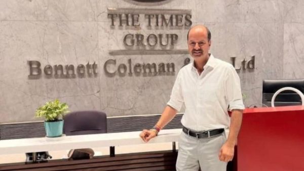 Man in a white shirt and gray pants stands at a reception desk in front of a marble wall with 'The Times Group Bennett Coleman & Co Ltd' sign, indoors with a small plant nearby.