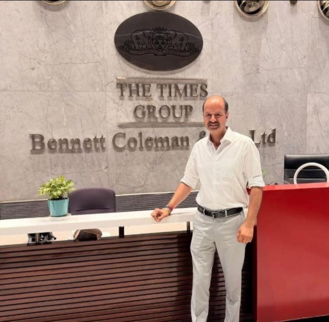 Man in a white shirt and gray pants stands at a reception desk in front of a marble wall with 'The Times Group Bennett Coleman & Co Ltd' sign, indoors with a small plant nearby.