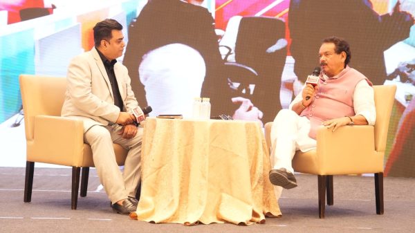 Two men sit in beige armchairs on stage, microphones in hand, during a talk show with a patterned table between them.