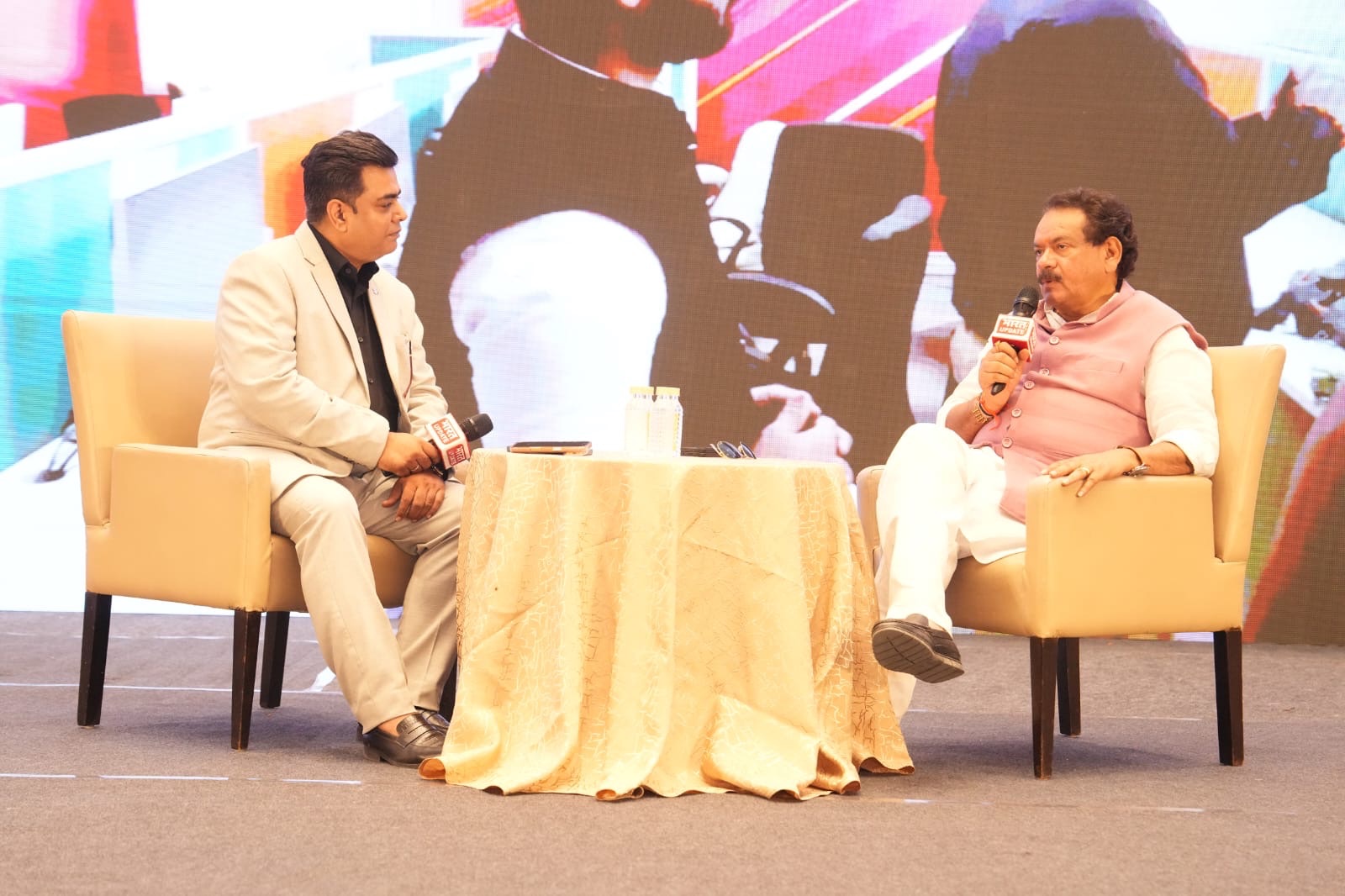 Two men sit in beige armchairs on stage, microphones in hand, during a talk show with a patterned table between them.