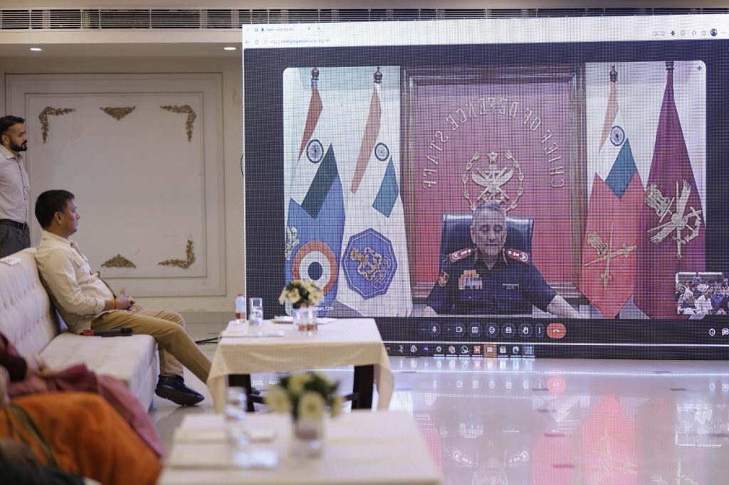 f8df1564-5ab4-450d-adbc-03c15f026087 | No. 1 Indian Media News Portal Man seated on a sofa watching a large video conference screen showing a police official at a desk with flags behind him.
